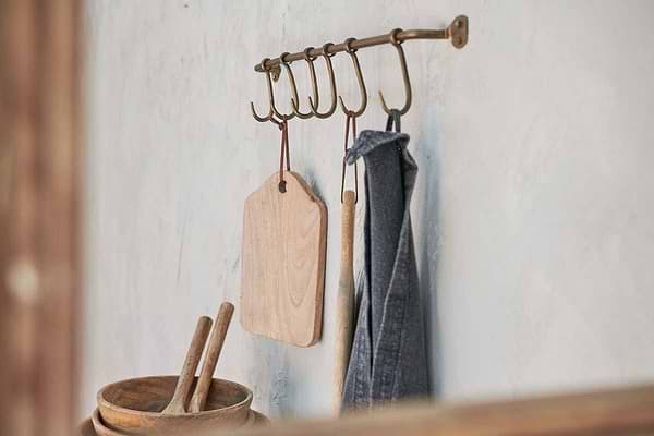 The nkuku Ikoma Hook Set in Antique Brass holds a wooden cutting board, spoon, and gray kitchen towel. A wooden bowl with utensils sits below, while the textured wall adds to the rustic, practical kitchen storage.