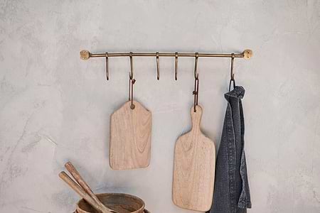 The nkuku Ikoma Hook Set - Antique Brass is mounted on a lightly textured wall, holding two wooden cutting boards and a dark towel. Below, a wooden bowl with utensils is partly visible in the corner.