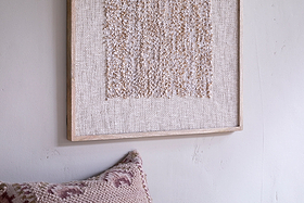 A close-up of nkuku's Ikona Textured Wall Art - Natural in a light wood frame hangs on a pale wall, with a blush pink and cream geometric pillow corner beneath, creating soft, neutral decor and an inviting feel.