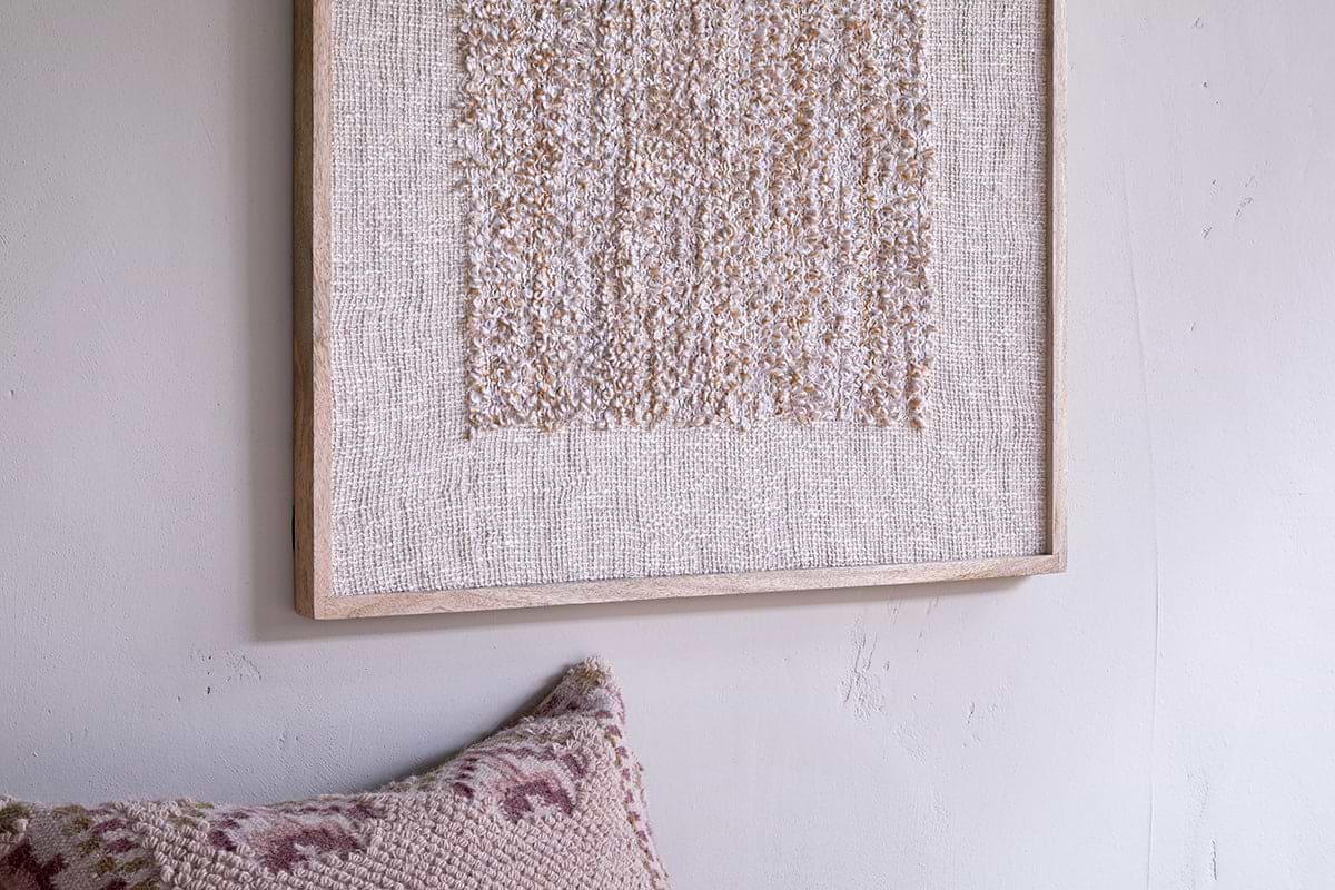 A close-up of nkuku's Ikona Textured Wall Art - Natural in a light wood frame hangs on a pale wall, with a blush pink and cream geometric pillow corner beneath, creating soft, neutral decor and an inviting feel.