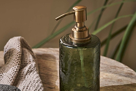 The Ilcoso Recycled Hammered Glass Soap Dispenser in green by nkuku rests on wood, with a textured beige towel nearby and soft green leaves in the background, creating a serene spa-like scene.