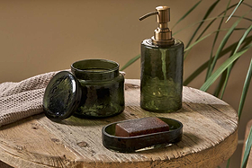 A rustic wooden table displays nkuku’s Ilcoso Recycled Hammered Glass Soap Dispenser in green, paired with matching lidded jar and soap dish. A beige towel and tall greenery enhance the natural, spa-inspired scene.