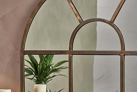 A close-up shows the nkuku Imoma Iron Overmantel Arch Mirror in a brass finish, reflecting a green plant in the lower left pane and bringing a natural touch to its elegant design.