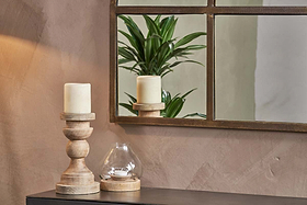 A beige wall features a black table topped with two wooden candle holders, cream candles, and a glass tealight holder. Above hangs the nkuku Imoma Iron Overmantel Arch Mirror in brass, reflecting green foliage in warm, cozy light.