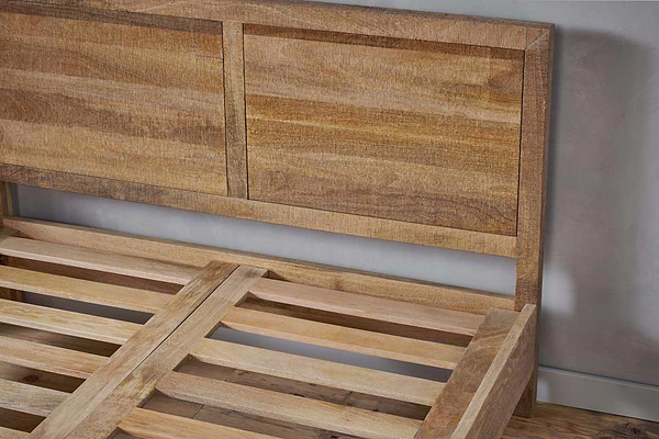 A close-up of the nkuku Indali Mango Wood Bed - Natural, featuring a light finish, two rectangular headboard panels, and a sturdy slatted base, shown against a gray wall and wooden floor.