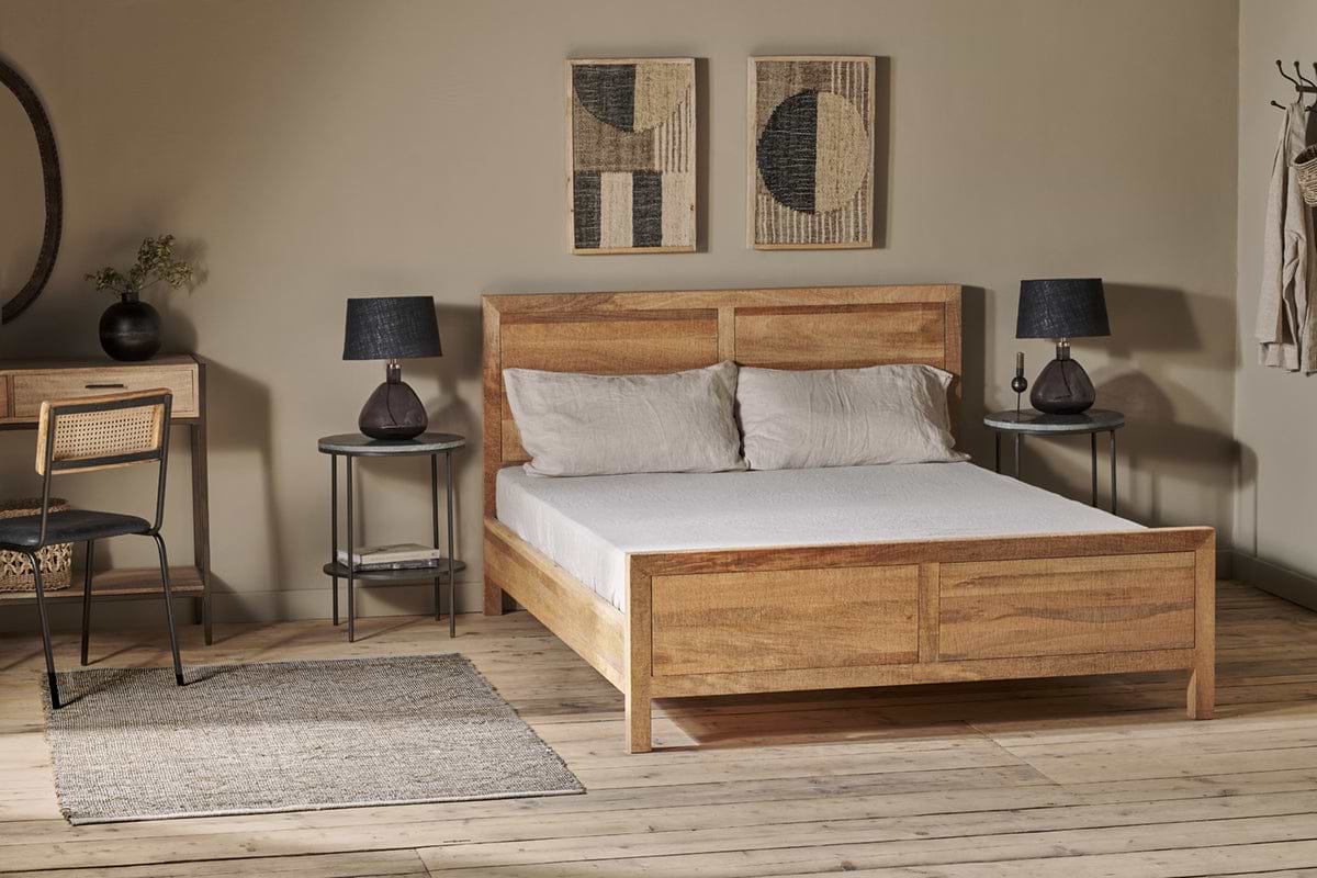 A cozy bedroom with the nkuku Indali Mango Wood Bed - Natural, two beige pillows, light gray bedspread, round side tables with black lamps, woven rug, small desk and chair, abstract art, and wooden flooring.