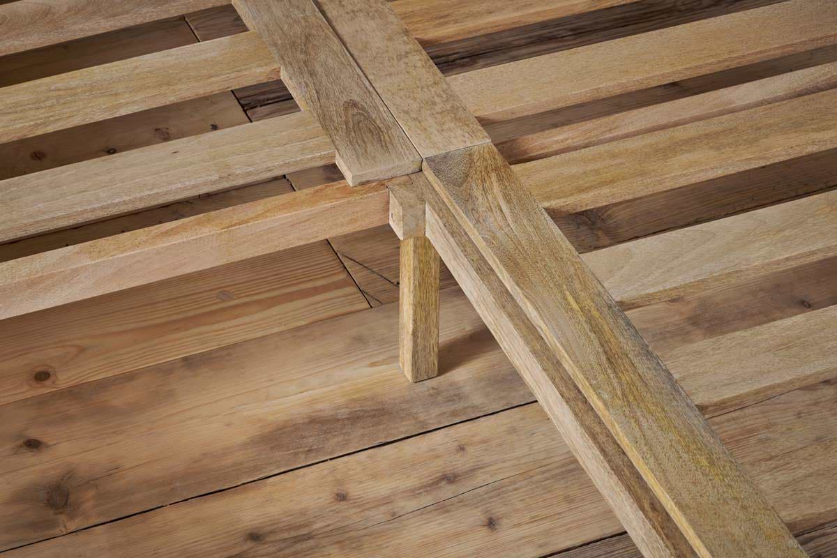 Close-up of the Indali Mango Wood Bed - Natural by nkuku shows sturdy, light mango wood slats and legs on a wooden floor. The smooth, unfinished wood highlights unique grain patterns and the bed’s natural texture.