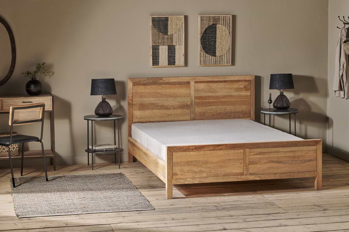 An Indali Mango Wood Bed - Natural by nkuku with white bedding sits center frame. Two black lamps with brown bases rest on grey side tables. Above, two abstract art pieces hang on a tan wall.
