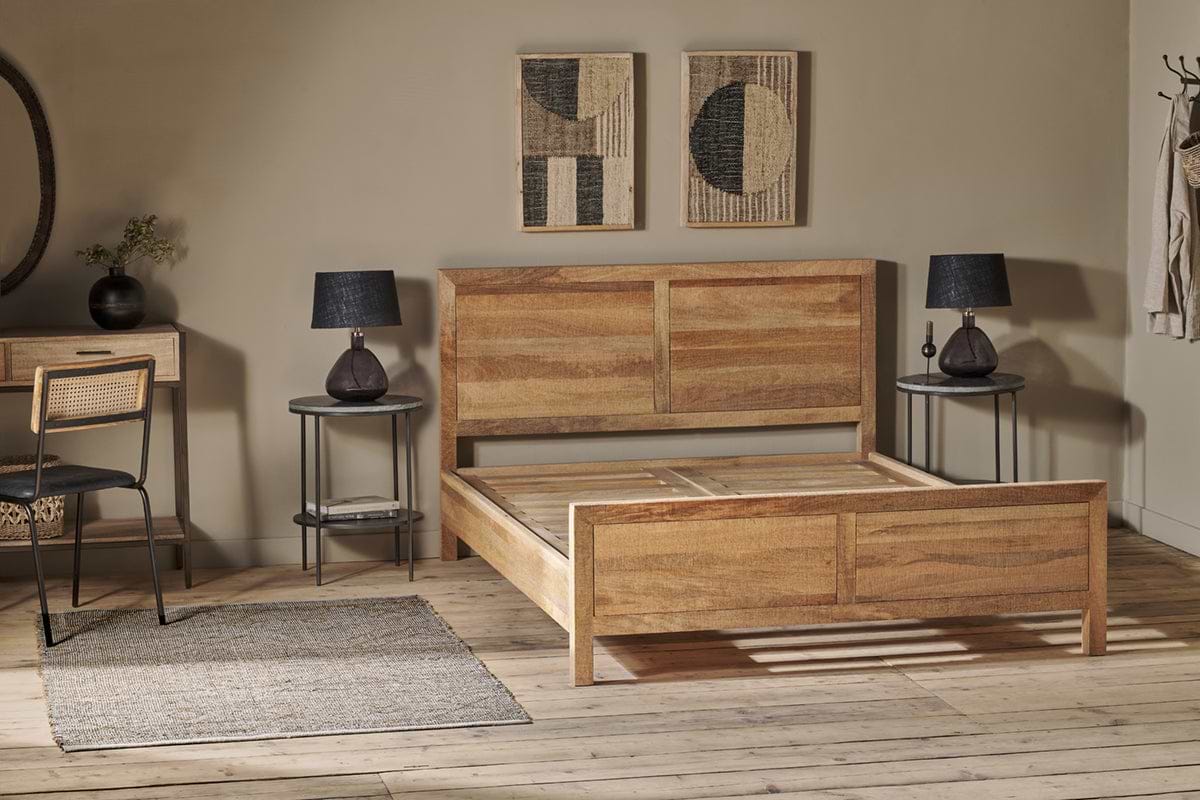 An Indali Mango Wood Bed - Natural by nkuku anchors a minimalist bedroom with light wood floors. Round nightstands with black lamps flank the bed, abstract art hangs above the headboard, and beige walls add a cozy, modern touch.