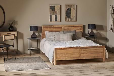 A cozy bedroom featuring the nkuku Indali Mango Wood Bed - Natural, white and beige bedding, two round black side tables with matching black lamps, a small desk and chair, a woven rug, wall art above the bed, and neutral wooden flooring.