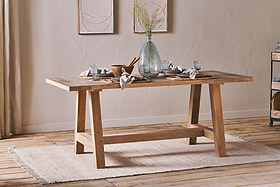The nkuku Indali Mango Wood Dining Table - Natural, with angled legs, stands on a beige rug in a cozy small space, set with glassware and a vase of dried flowers beneath neutral wall art as sunlight streams through the curtains.