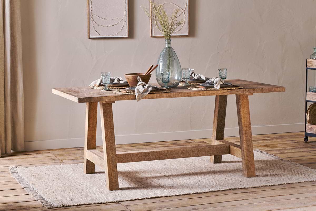 The nkuku Indali Mango Wood Dining Table - Natural, with angled legs, stands on a beige rug in a cozy small space, set with glassware and a vase of dried flowers beneath neutral wall art as sunlight streams through the curtains.