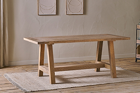 The Indali Mango Wood Dining Table - Natural by nkuku, with angled legs, sits on a beige textured rug in a small space with light wood floors, neutral walls, and two abstract artworks softly lit by natural light.