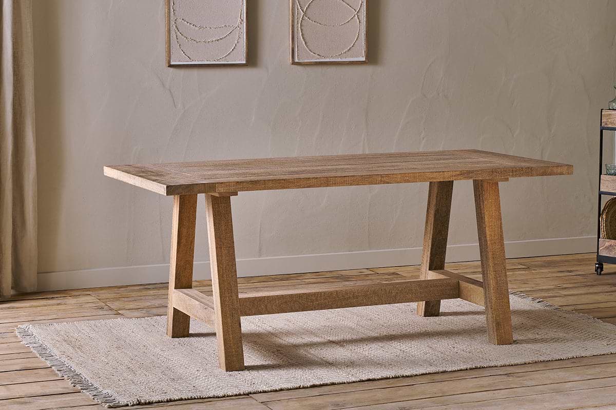 The Indali Mango Wood Dining Table - Natural by nkuku, with angled legs, sits on a beige textured rug in a small space with light wood floors, neutral walls, and two abstract artworks softly lit by natural light.