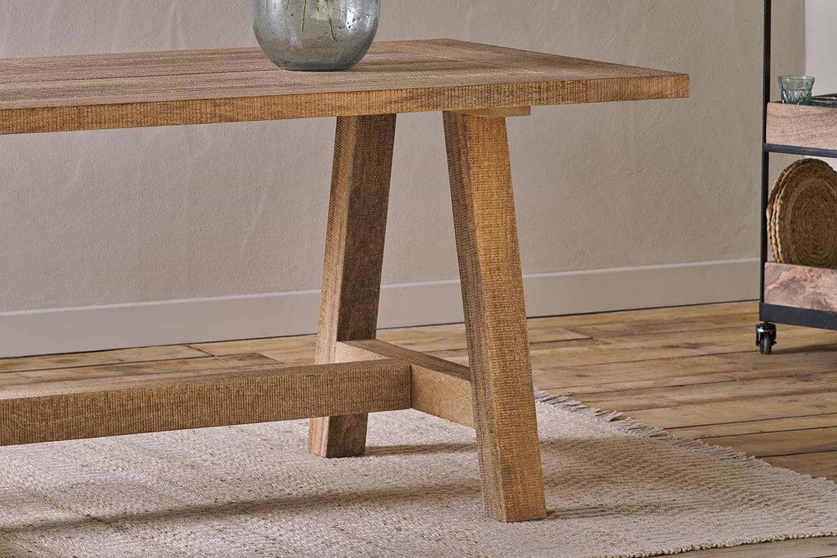 A close-up of the nkuku Indali Mango Wood Dining Table - Natural, featuring angled legs on a textured beige rug. Perfect for small spaces, it displays a rustic finish with a ceramic vase, wooden floor, and metal shelves in the background.