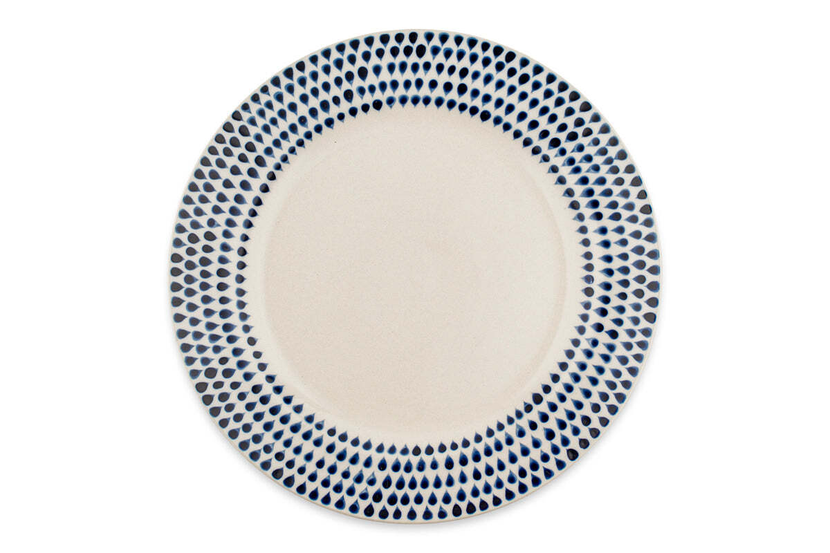 The Indigo Drop Dinnerware Set - Cream & Indigo (Set of 12) by nkuku features round, handmade ceramic plates from Vietnam with hand-painted indigo teardrop rims and plain centers that emphasize the bold border design.