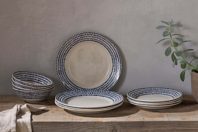 A rustic wooden table displays the nkuku Indigo Drop Dinnerware Set - Cream & Indigo (Set of 12), featuring handmade plates and bowls with blue dotted rims. A beige cloth sits to the left, beside a potted plant against a neutral gray wall.