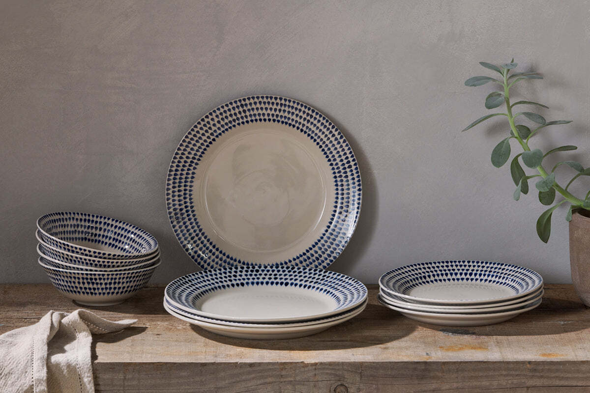 A rustic wooden table displays the nkuku Indigo Drop Dinnerware Set - Cream & Indigo (Set of 12), featuring handmade plates and bowls with blue dotted rims. A beige cloth sits to the left, beside a potted plant against a neutral gray wall.