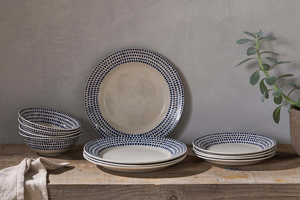 A rustic wooden table displays the nkuku Indigo Drop Dinnerware Set - Cream & Indigo (Set of 12), featuring handmade plates and bowls with blue dotted rims. A beige cloth sits to the left, beside a potted plant against a neutral gray wall.