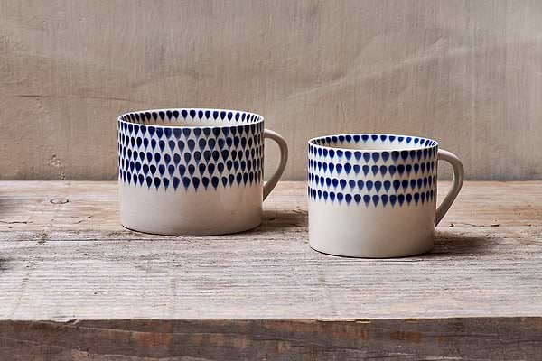The nkuku Indigo Drop Mug - Cream & Indigo - Large (Set of 2) features handmade ceramic mugs with indigo teardrop patterns near the rim, displayed on a rustic wooden surface against a neutral textured background.