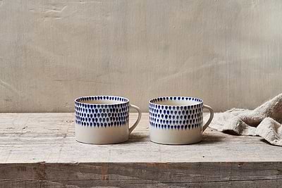 The nkuku Indigo Drop Mug - Cream & Indigo - Large (Set of 2) features handmade ceramic mugs with white bases and blue teardrop designs, displayed side by side on rustic wood with a beige textured cloth and a light background.