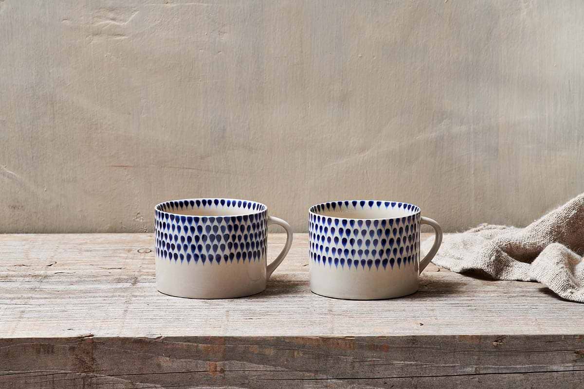 The nkuku Indigo Drop Mug - Cream & Indigo - Large (Set of 2) features handmade ceramic mugs with white bases and blue teardrop designs, displayed side by side on rustic wood with a beige textured cloth and a light background.