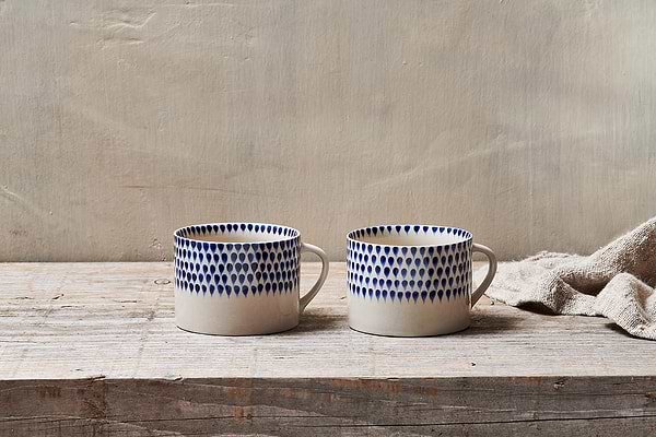 The nkuku Indigo Drop Mug - Cream & Indigo - Large (Set of 2) features handmade ceramic mugs with white bases and blue teardrop designs, displayed side by side on rustic wood with a beige textured cloth and a light background.