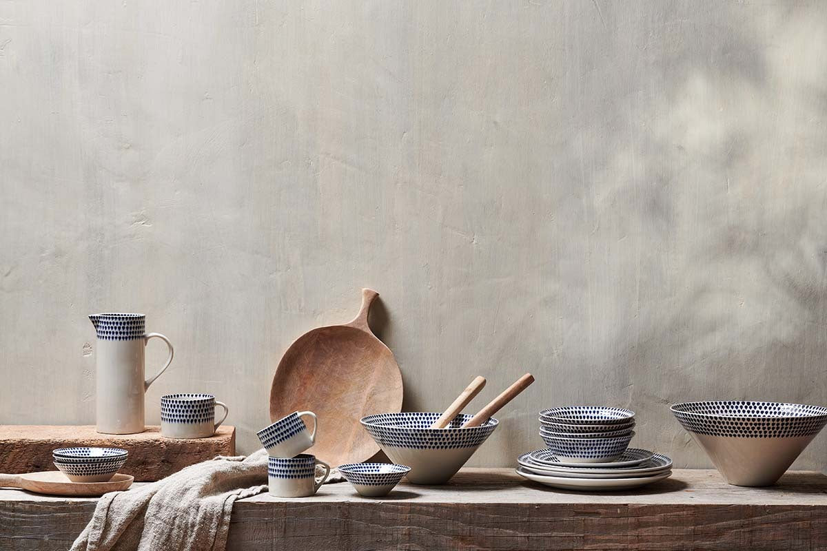 A rustic wooden surface displays nkuku Indigo Drop Mugs in cream and indigo (small, set of 2) with blue dotted patterns. Wooden utensils rest in a bowl; a wooden tray and beige cloth add texture against a light, textured wall.