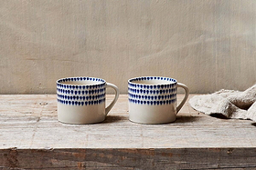 The nkuku Indigo Drop Mug - Cream & Indigo - Small (Set of 2) features hand-painted indigo designs on white ceramic, displayed side by side on rustic wood with a beige cloth for a minimalist, cozy look.
