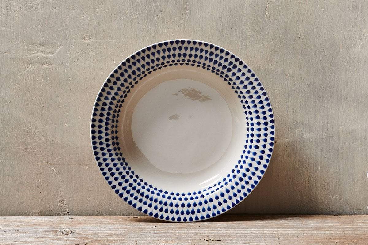 The Indigo Drop Pasta Bowl - Cream & Indigo by nkuku, featuring handpainted indigo dots on off-white ceramic, rests on wood and pops against a textured beige wall.