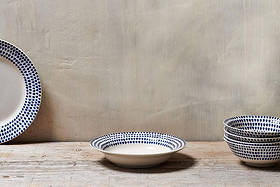 On a rustic wooden surface, the nkuku Indigo Drop Pasta Bowl - Cream & Indigo is displayed alongside handpainted bowls, their blue dotted patterns creating a charming, artisanal look against a plain beige wall.