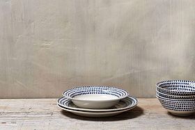 Three handmade ceramic plates with blue dotted patterns are stacked on a rustic wooden surface, alongside four nkuku Indigo Drop Pasta Bowls in Cream & Indigo. A plain beige textured wall forms the minimalist background.