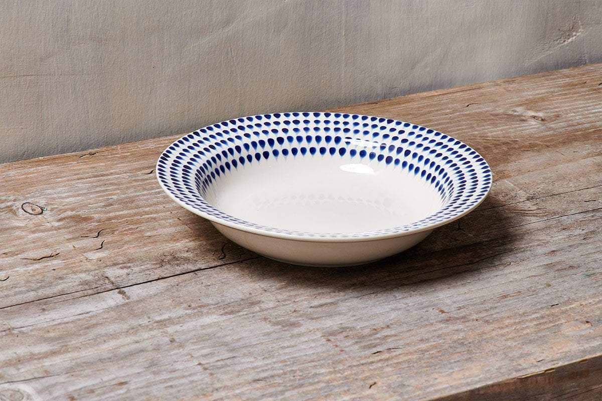 The nkuku Indigo Drop Pasta Bowl - Cream & Indigo, handmade with a blue dotted rim on cream ceramic, sits empty and centered on a rustic wooden surface against a light beige wall.