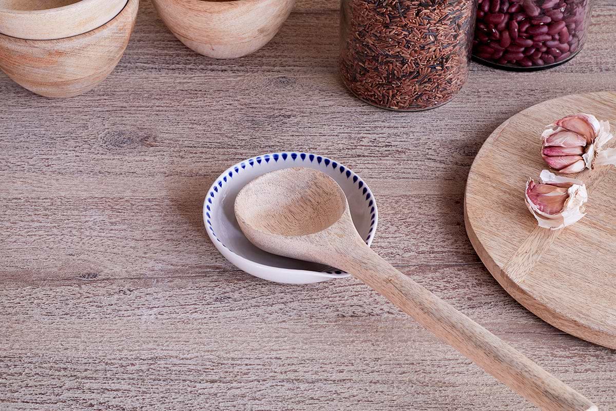 A wooden spoon sits on the nkuku Indigo Drop Spoon Rest - Cream & Indigo, placed in a small white dish with blue dots. Around it are handcrafted wooden bowls, jars of rice and beans, and a cutting board with garlic for a rustic kitchen vibe.