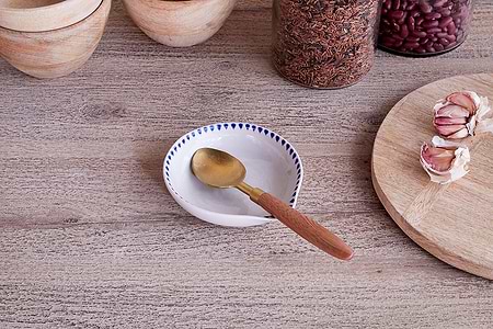 The nkuku Indigo Drop Spoon Rest in cream & indigo sits on a wooden surface, holding a brass spoon with a wooden handle. Nearby are stacked wooden bowls, jars of rice and beans, a wooden board, and cloves of garlic.