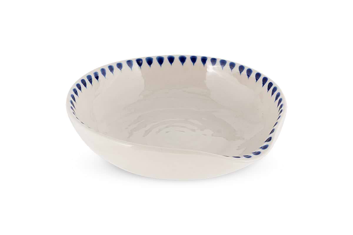 The Indigo Drop Spoon Rest - Cream & Indigo by nkuku is a shallow, round handcrafted bowl with an off-white glaze, uneven rim, and blue teardrop ceramic edging, ideal as a decorative spoon rest. Plain white background.