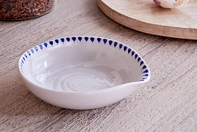 The nkuku Indigo Drop Spoon Rest in cream and indigo, featuring blue teardrop patterns, rests on a light wooden surface with a round wooden board and a jar of red rice in the background.