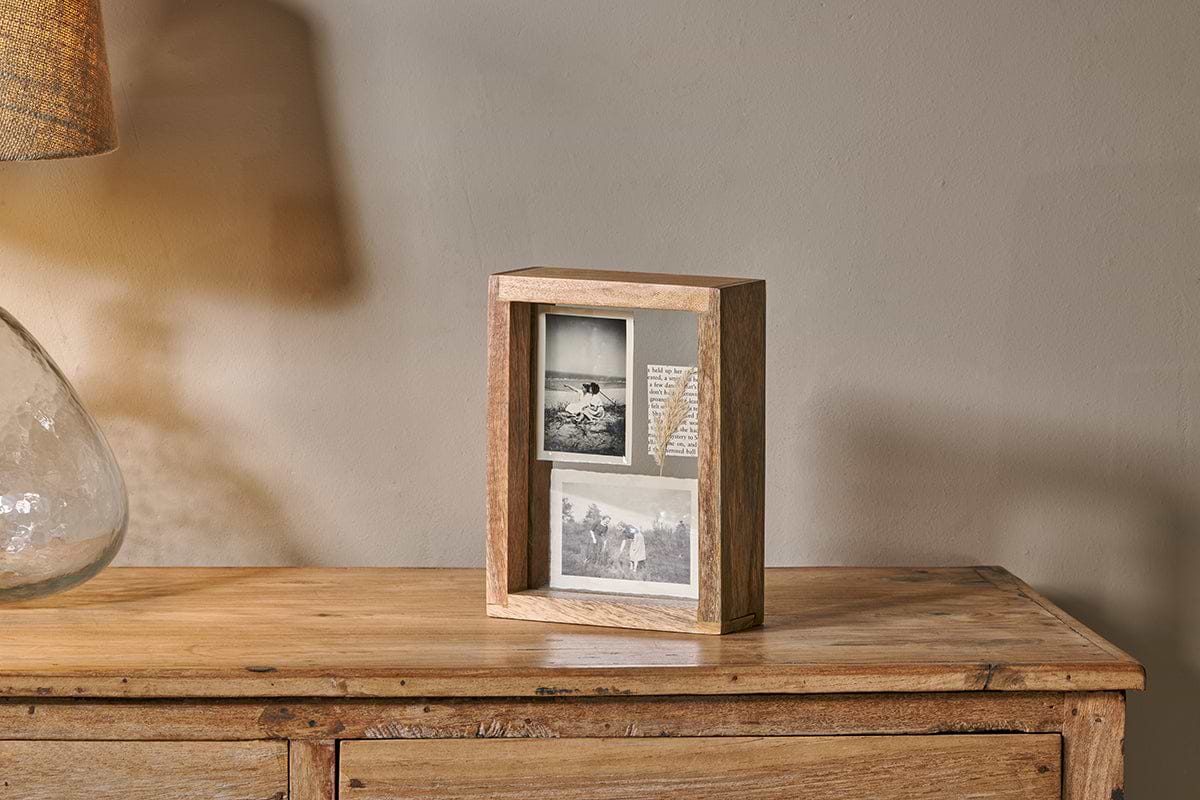 The nkuku Indu Standing Mango Wood Frame - Natural holds two black-and-white photos and text on a rustic dresser, with a glass lamp and beige shade partially visible to the left against a plain light wall.