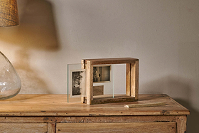 The Indu Standing Mango Wood Frame - Natural by nkuku, made from sustainable mango wood, holds two black-and-white photos on a wooden table with a glass lamp. Warm light highlights the frame near a plain beige wall, chalk resting nearby.