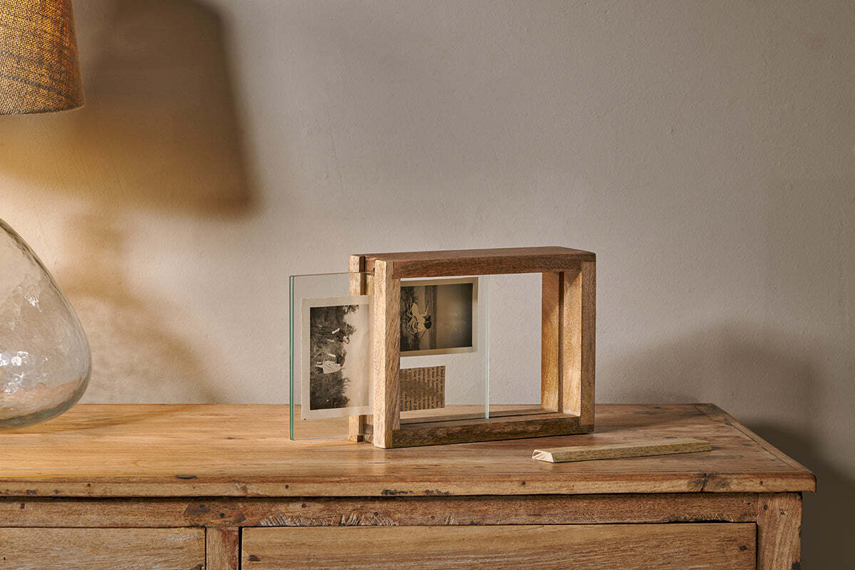 The Indu Standing Mango Wood Frame - Natural by nkuku, made from sustainable mango wood, holds two black-and-white photos on a wooden table with a glass lamp. Warm light highlights the frame near a plain beige wall, chalk resting nearby.