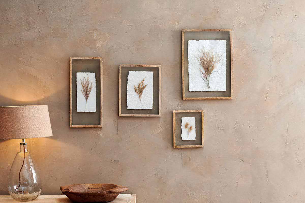 Four botanical artworks, each showcased in the Indu Wooden Frame made from solid mango wood by nkuku, display dried plants on white backgrounds against a beige textured wall. The frames' natural mango wood highlights the organic textures of the art. Beneath the arrangement, a clear glass lamp with a tan shade and a carved wooden bowl rest on a light wood table.