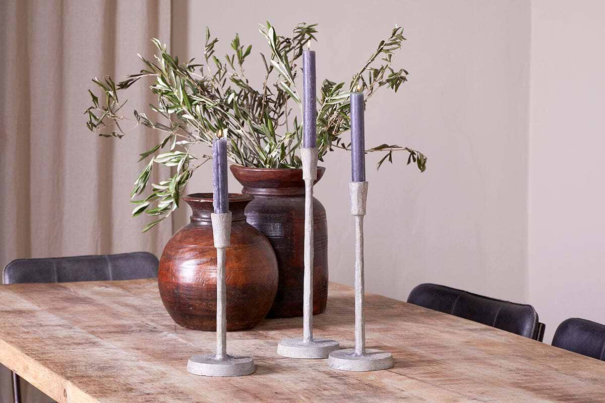 Three aged zinc Inigo Candle Holders by nkuku, each holding a purple candle, stand on a rustic wooden table. Behind them are two round, brown vases filled with leafy green branches. Black chairs surround the table, and beige curtains are visible in the background.
