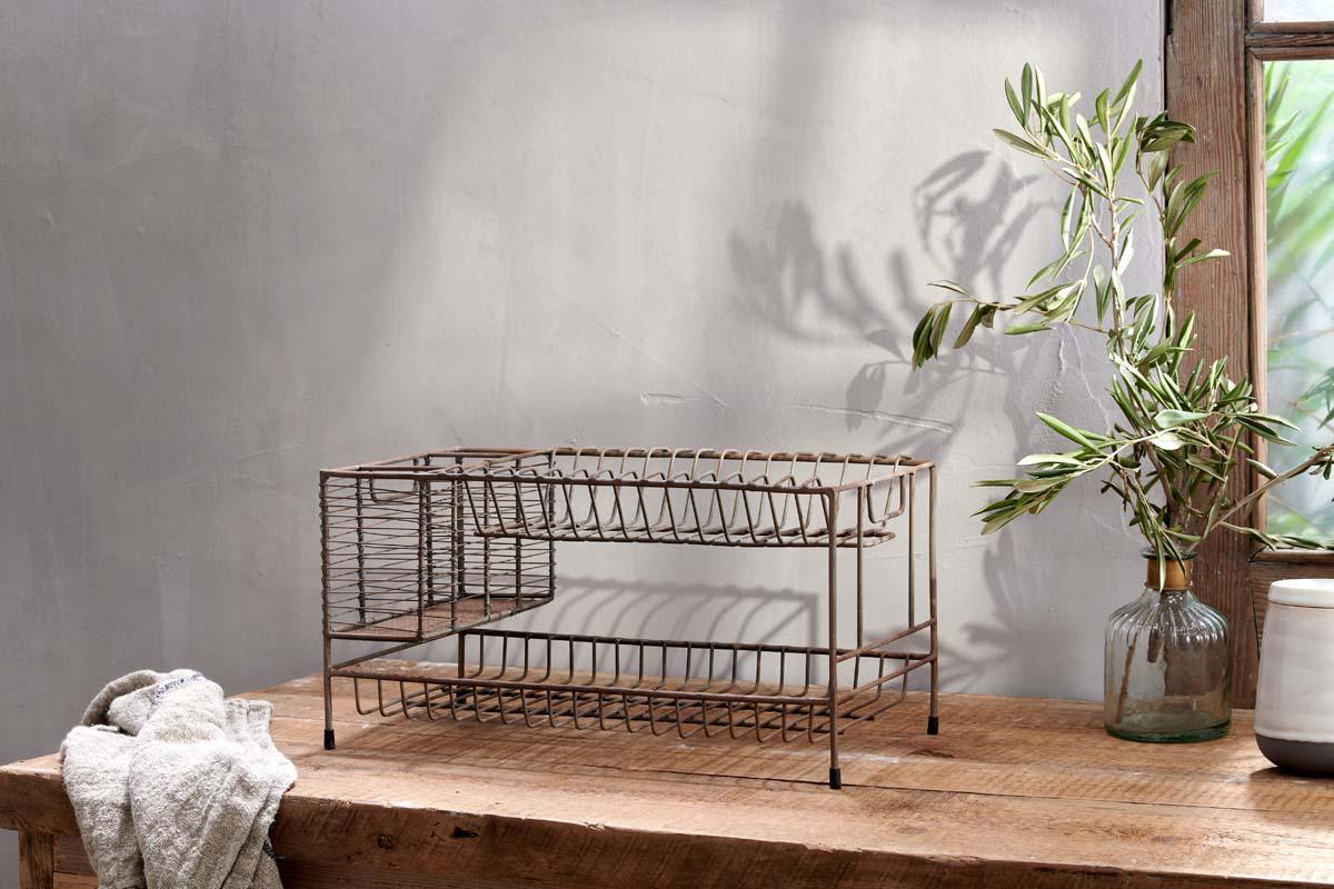 A rustic wooden table displays nkuku’s Inkollu Dish Rack in aged brass, a clear vase with olive branches, a white cup, and a light towel. Soft daylight casts gentle shadows on the gray wall, creating a calm, natural mood.