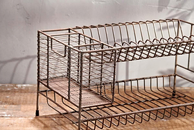 The Inkollu Dish Rack by nkuku, a vintage two-tiered design with wire compartments and an aged brass finish, sits on a wooden surface against a textured light gray wall, showcasing rustic charm and visible wear.