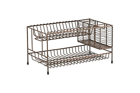 The nkuku Inkollu Dish Rack features two metal tiers with an aged brass finish. The top slots hold plates, the lower level fits larger items, and a side wire basket stores utensils. Four black-tipped feet enhance its utilitarian design.