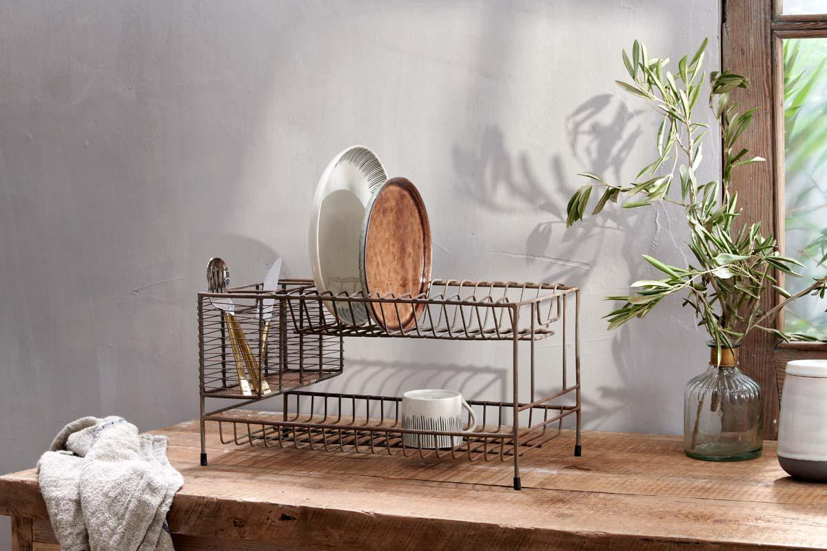 The nkuku Inkollu Dish Rack sits on a wooden counter, organizing plates, cutlery, and a mug. A beige towel and brass vase with green leaves nearby catch sunlight by the window, casting gentle shadows on the gray wall.