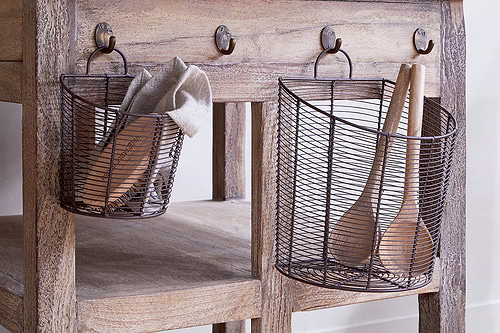 The nkuku Inkollu Wall Hung Baskets in aged brass (set of 2) hang from hooks on a rustic wooden table with a natural, weathered finish; one basket holds a beige cloth and utensil, the other two wooden spoons.