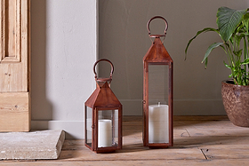 Two nkuku Isko Lanterns in a rich rust finish, each with glass panels and a white pillar candle, stand on wooden flooring near a beige wall. A green potted plant and wooden door add charm—perfect for cozy garden seating areas.
