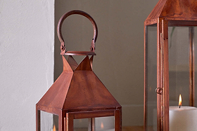 A close-up of two Isko Lanterns - Rust by nkuku. These rustic, copper-toned lanterns feature a rust finish, glass panels, and white pillar candles. They rest on a wooden floor near a white wall, ideal for cozy garden seating.