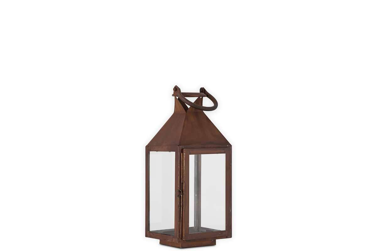 The Isko Lantern - Rust by nkuku features a vintage-style rectangular frame with clear glass panels, a rust finish, pointed top, metal loop handle, and flat base—ideal for pillar candles or adding rustic charm to your garden seating.